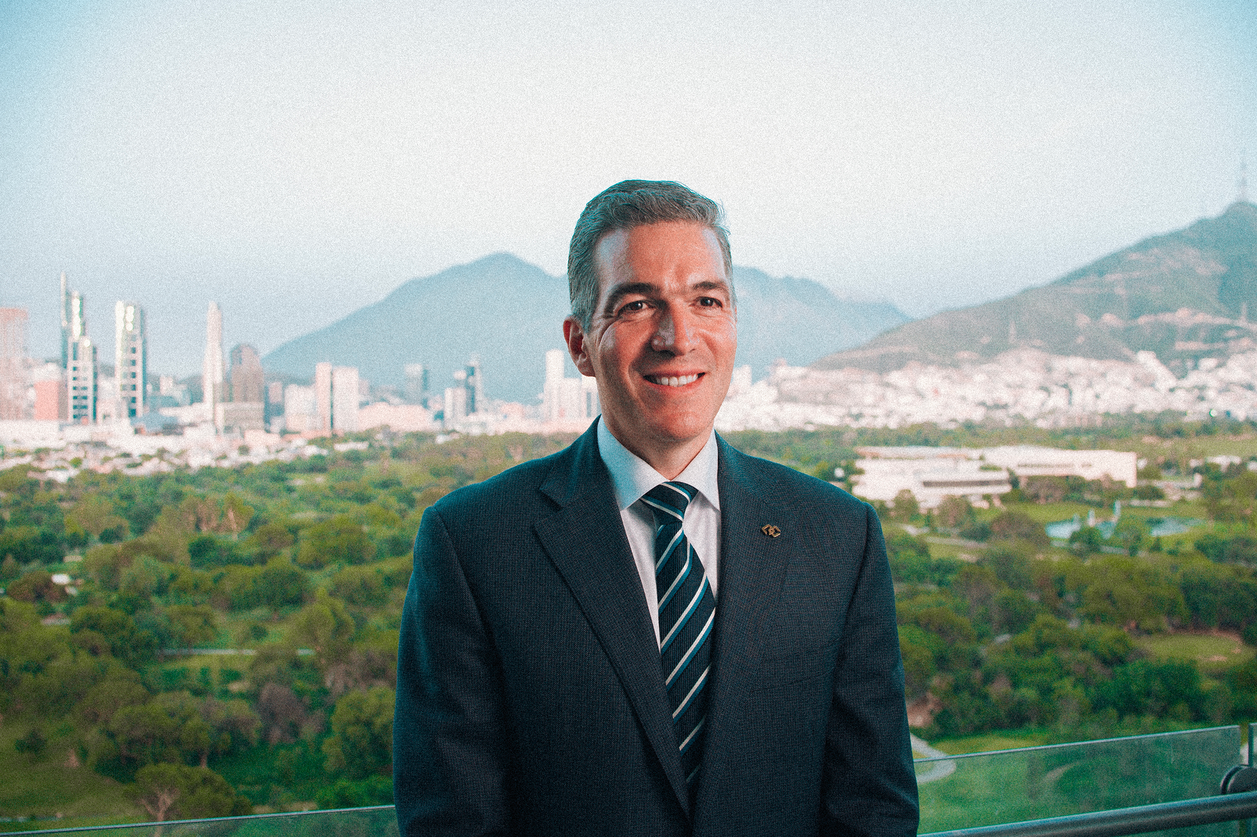 Un hombre con traje se encuentra en un balcón con vistas a un paisaje urbano con edificios altos, un parque verde y montañas al fondo.